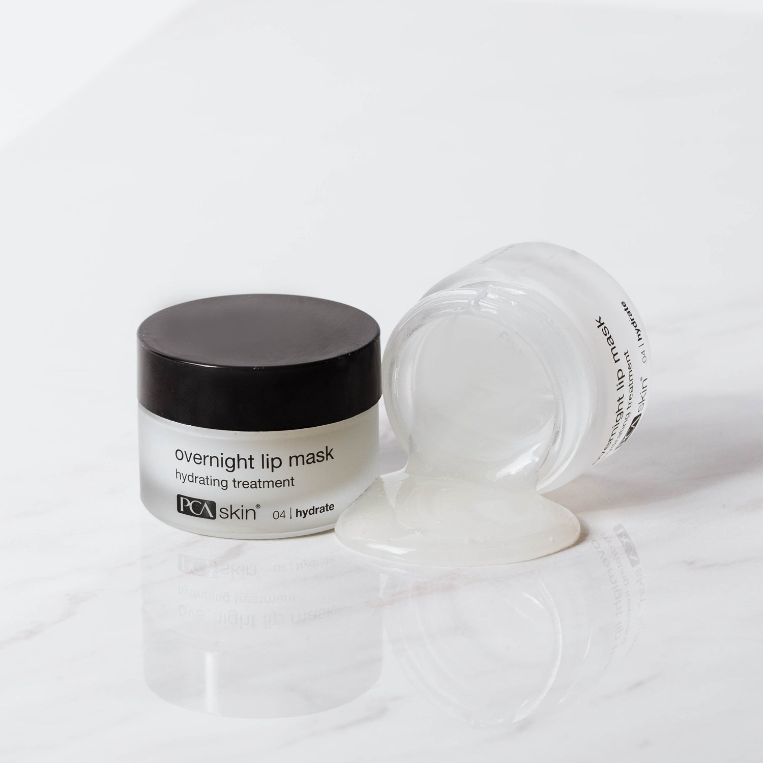 Image of an overnight lip mask by PCA Skin. The product, which comes in a small jar with a black lid, is labeled "HA Lip Overnight Mask." The jar is shown both closed and open, with some of the clear gel-like mask oozing out onto a white marble surface.