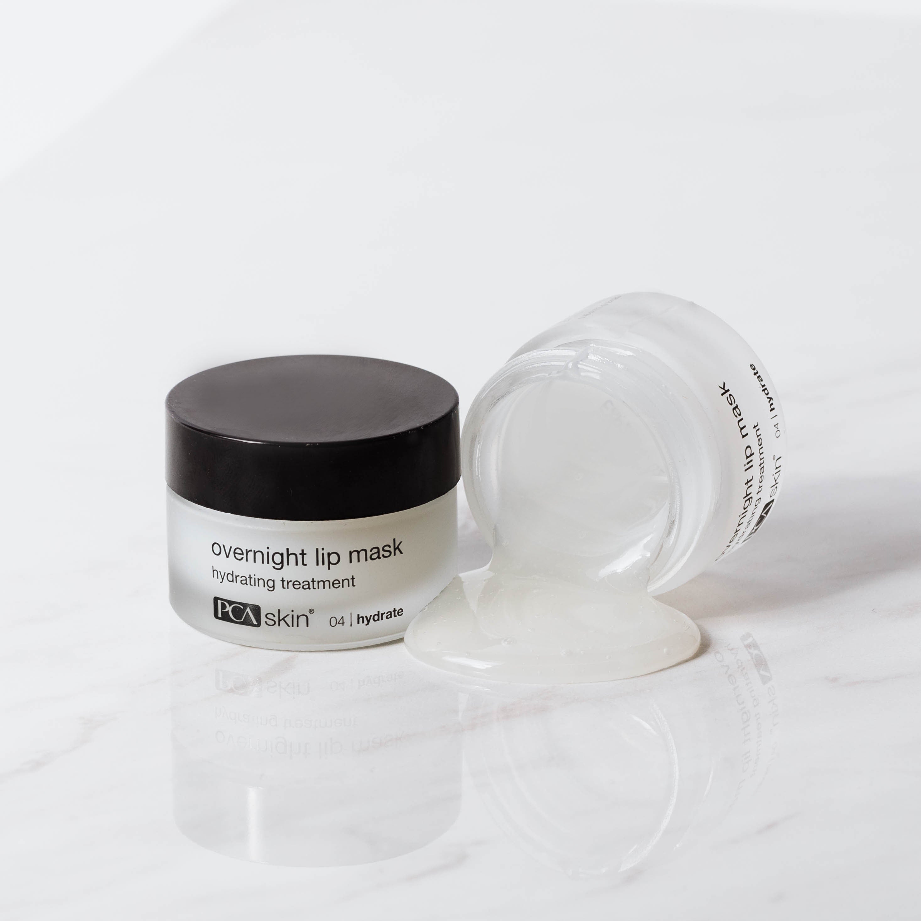 Image of an overnight lip mask by PCA Skin. The product, which comes in a small jar with a black lid, is labeled "HA Lip Overnight Mask." The jar is shown both closed and open, with some of the clear gel-like mask oozing out onto a white marble surface.