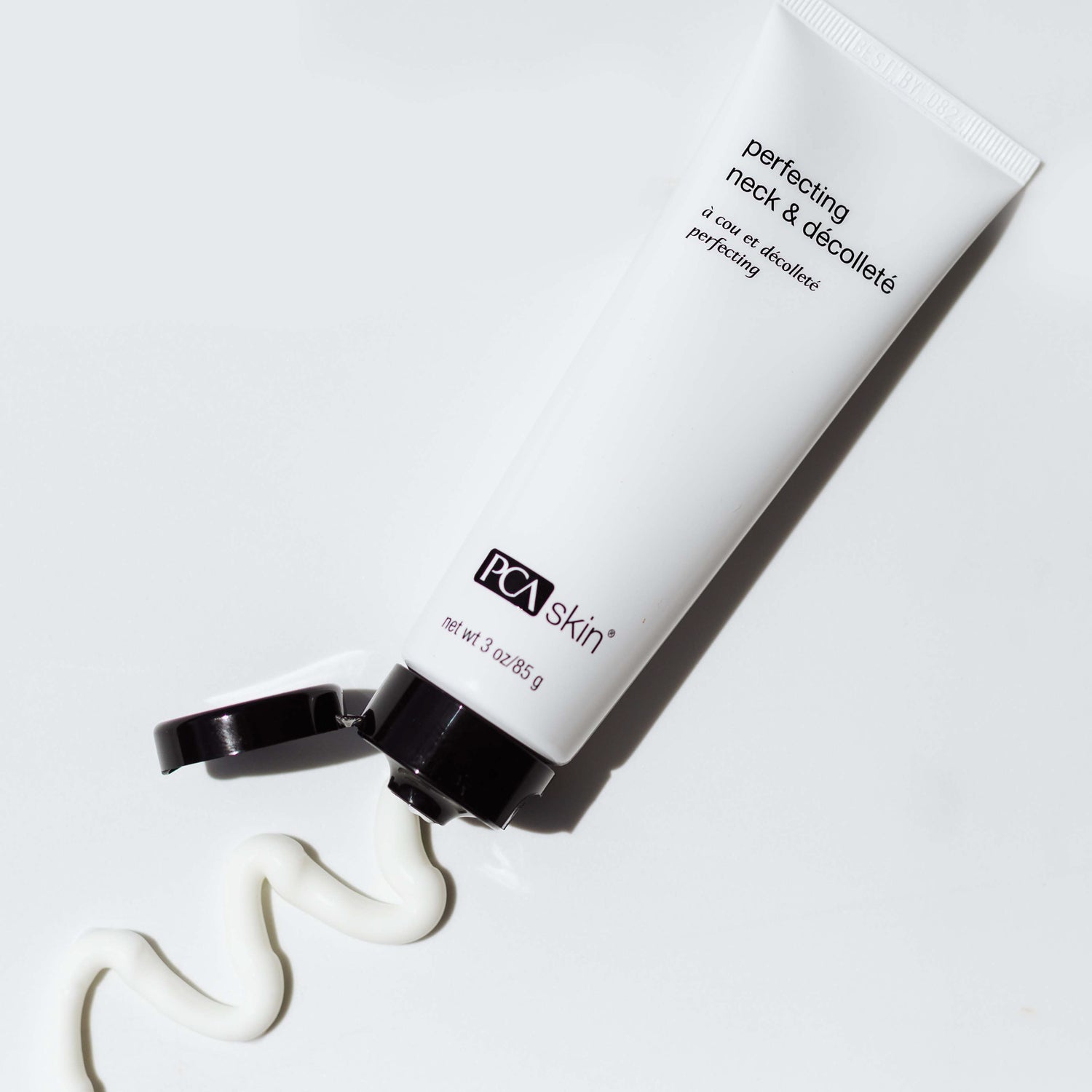 A white tube of Perfecting Neck & Décolleté lies on its side with the cap open, dispensing a wavy line of cream onto a white surface. The tube is labeled with brand and product information, promising to address uneven skin tone and fine lines.