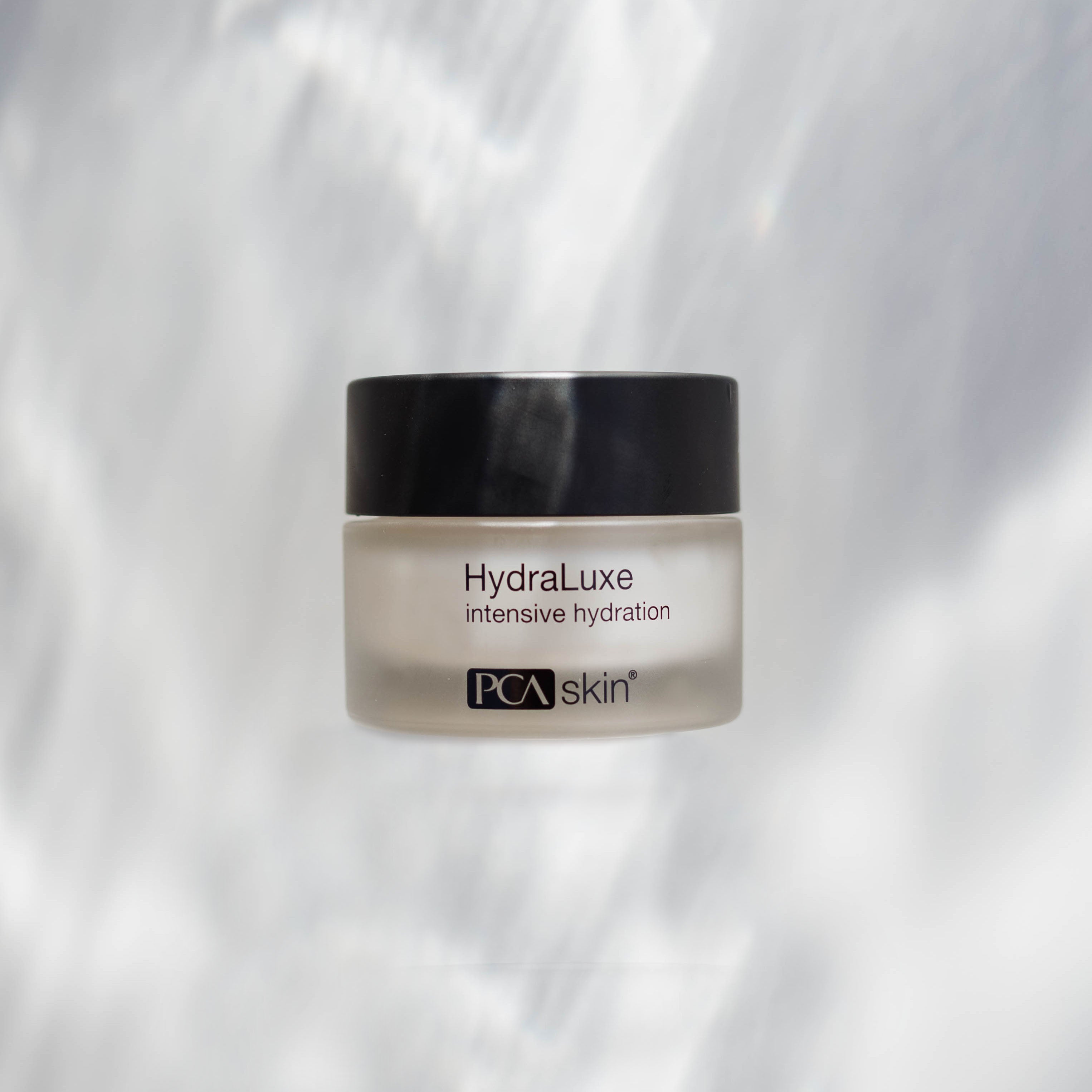 A frosted glass jar with a black lid containing HydraLuxe Intensive Hydration cream. The text on the jar reads "HydraLuxe intensive hydration." Providing deep hydration, this cream also softens fine lines. The background is light with subtle blurred patterns.