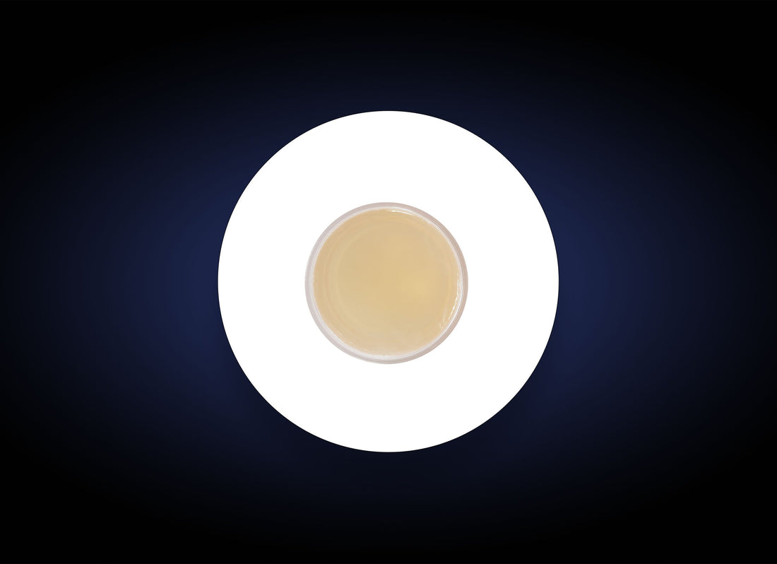 A top-down view of a cup filled with a light beige liquid, possibly tea or coffee, placed on a white circle against a dark background. The contrast between the white circle and dark backdrop highlights the cup centrally, akin to how the Blemish Control Bar targets acne breakouts on oily skin.