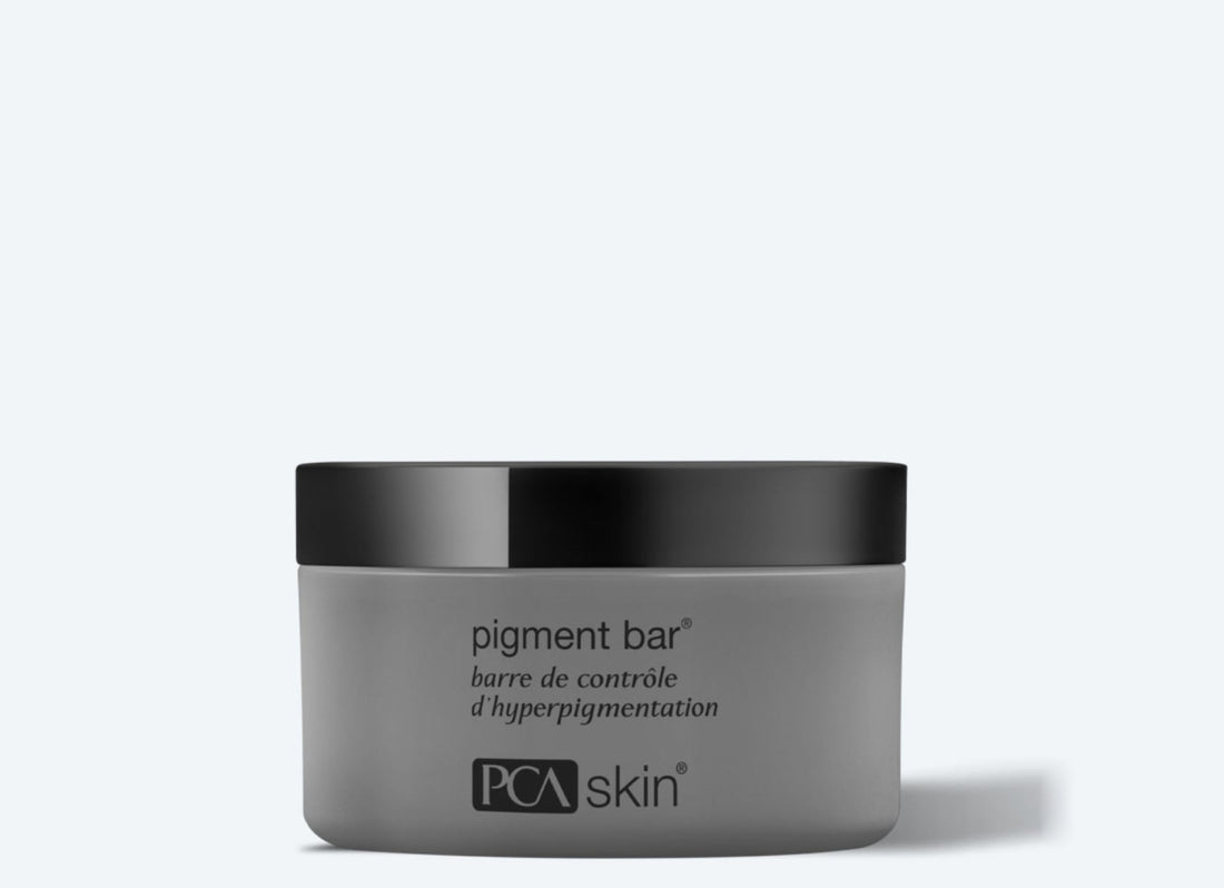 A gray jar of Pigment Bar with a black lid, designed for hyperpigmentation control and even skin tone. The text on the jar reads "pigment bar" and "barre de contrôle d’hyperpigmentation" above the "PCA skin" logo at the bottom. The background is light gray.