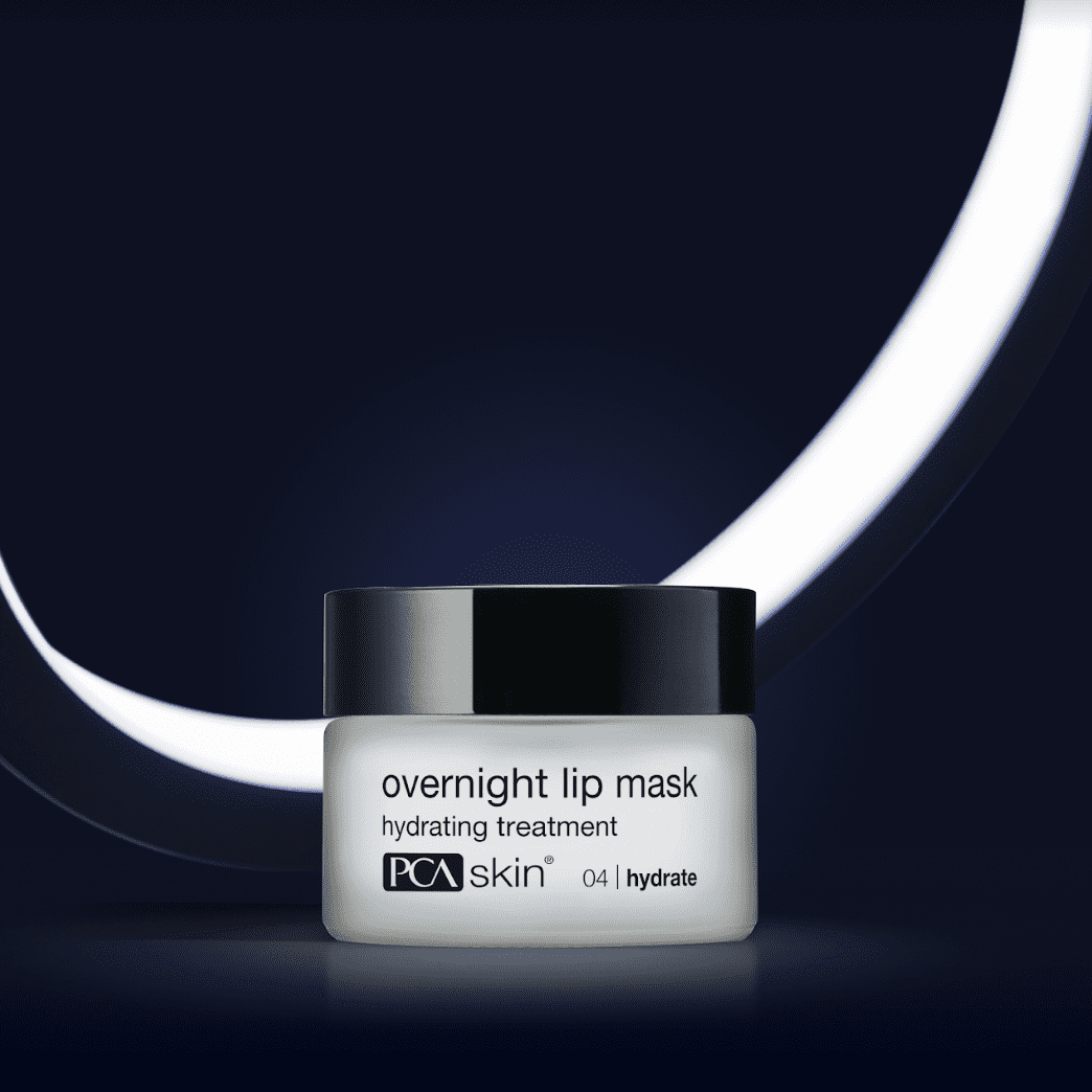 A white jar with a black lid labeled "HA Lip Overnight Mask" from PCA Skin is in the center of a dark background with an illuminated white circular light partially visible behind the jar, creating a sleek and modern look. This ultra-hydrating nighttime lip treatment fights visible signs of ageing.