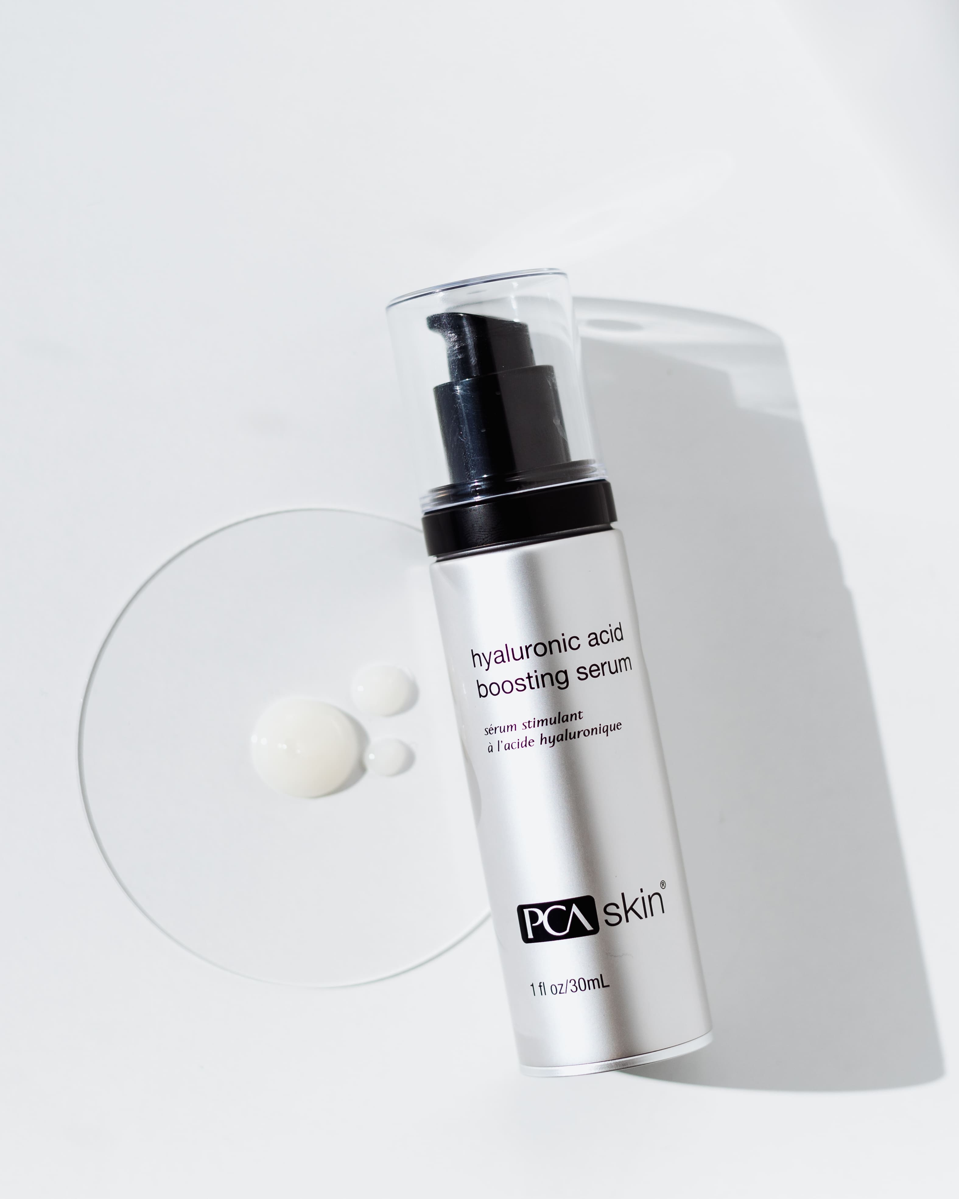 A 1 fl oz bottle of Hyaluronic Acid Boosting Serum with a pump dispenser is shown against a white background. Some of the serum, known for its powerful hydration and anti-aging properties, is dispensed next to the bottle, displaying its clear and creamy texture. The bottle has a sleek black and white design.