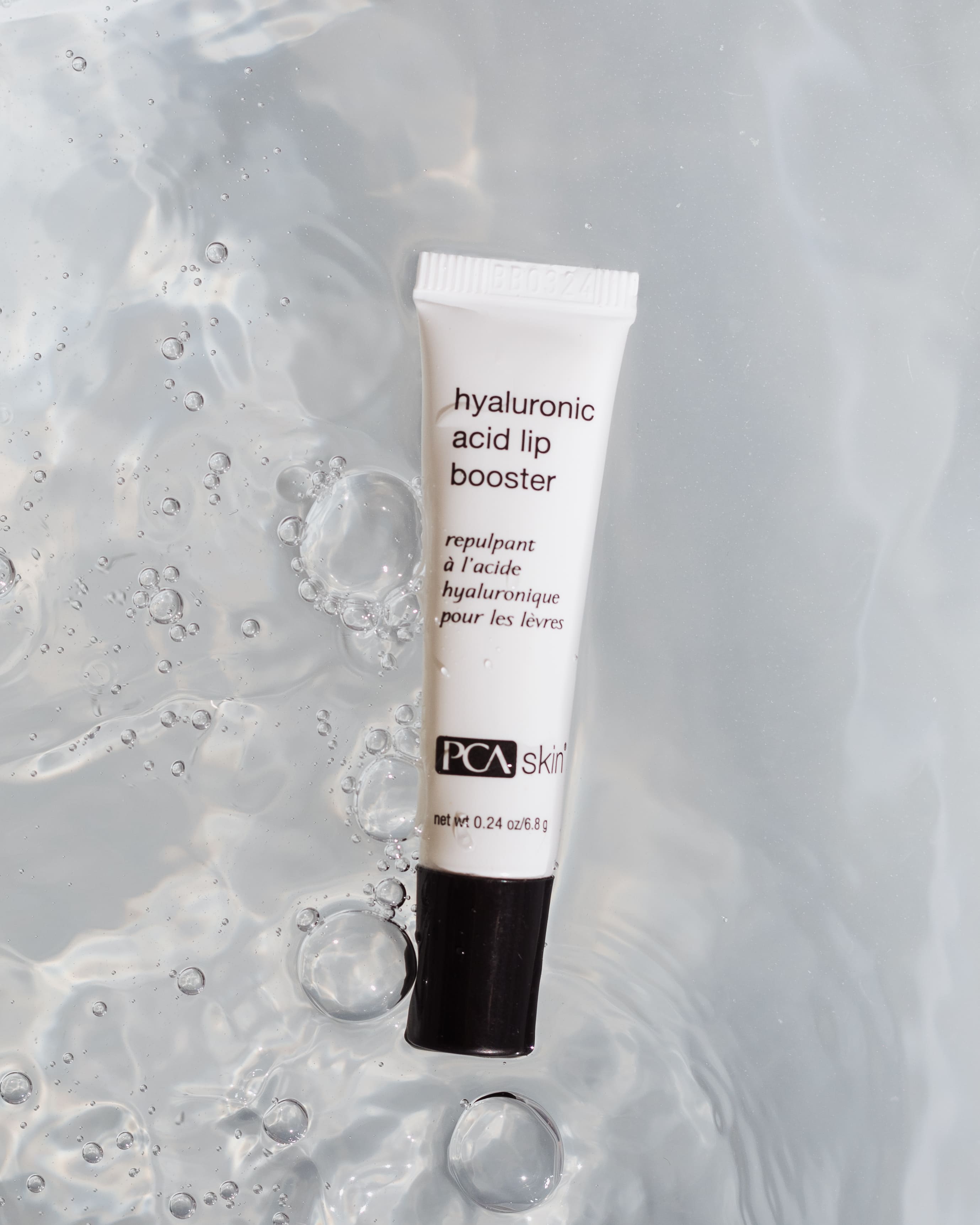 A tube of Hyaluronic Acid Lip Booster on a wet, reflective surface with scattered water droplets around it. The tube is white with black text and has the brand "PCA Skin" printed on the bottom, promising to enhance your lips while supporting collagen production.