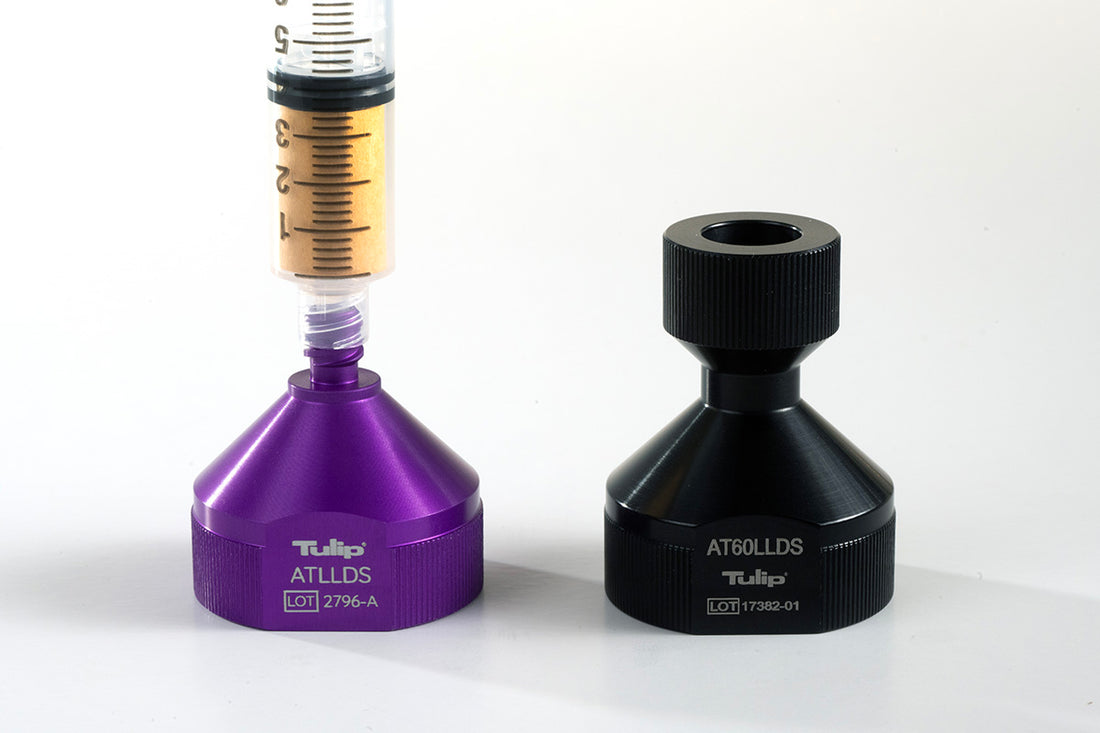 A close-up of two medical devices on a white background. The Decanting Stand™ on the left is purple and labeled "Tulip, ATLLDS, LOT 2796-A," with a syringe inserted at the top, designed for anaerobic transfers. The Decanting Stand™ on the right is black and labeled "AT60LLDS, LOT 17382-01.