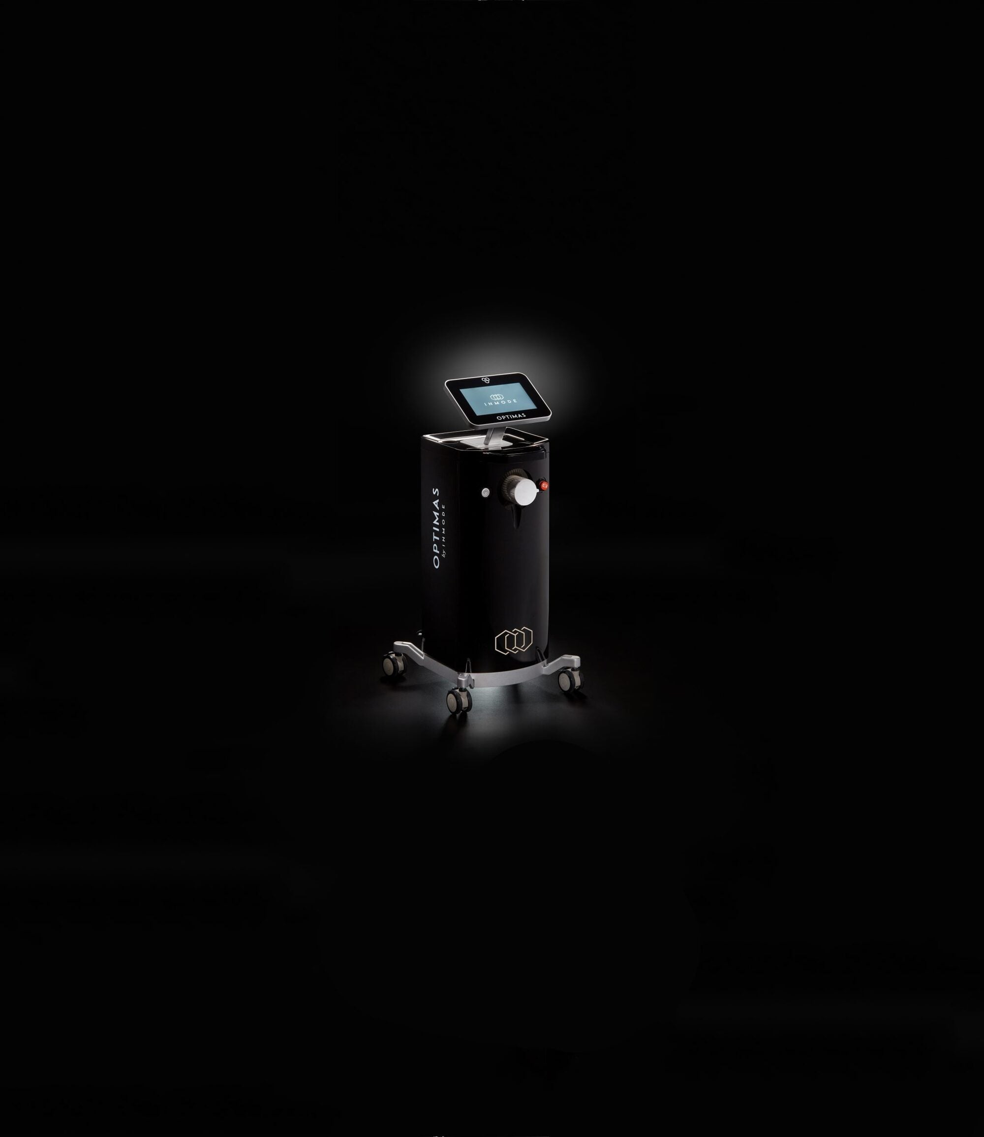 A sleek, modern floor-standing Optimas coffee machine on wheels illuminated by a spotlight against a dark background. The Optimas features a touch screen interface and is part of a full beauty suite, complete with a coffee cup dispenser.