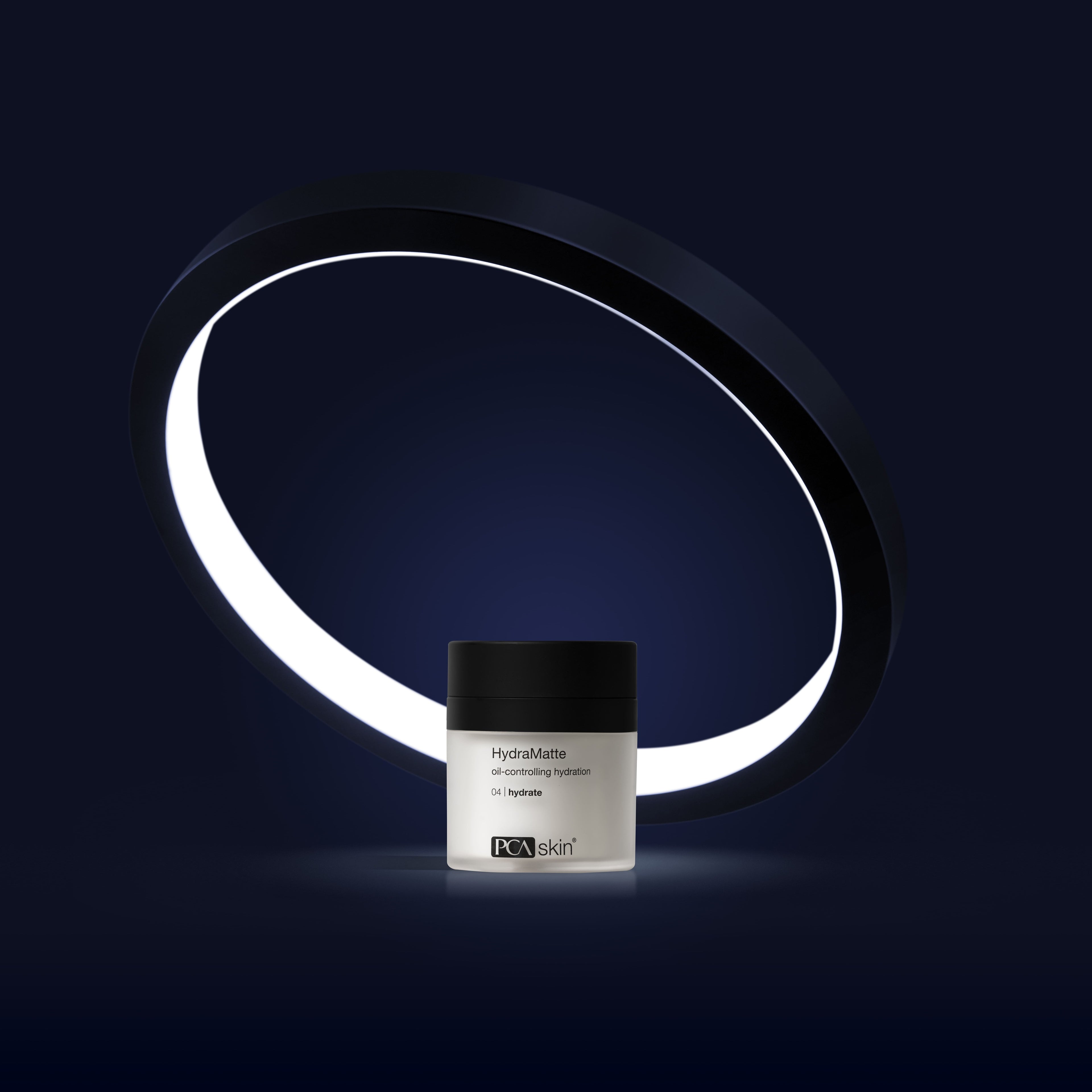 A small, white jar with a black lid labeled &quot;HydraMatte&quot; sits against a dark background. The jar is centered within a large, illuminated circular ring that glows brightly, emphasizing the HydraMatte&