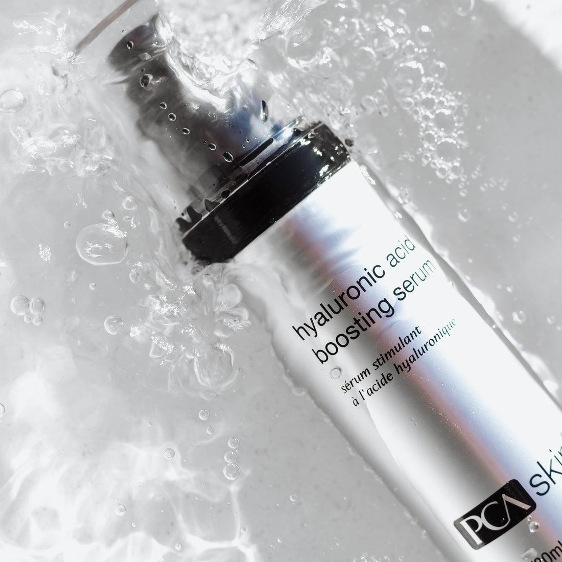 A gray bottle of **Hylauronic Acid Boosting Serum**, known for its anti-aging and hydration benefits, lies on a white surface splashed with water. The bottle features a gradient design, transitioning from metallic to white, with black text displaying the product name and brand.