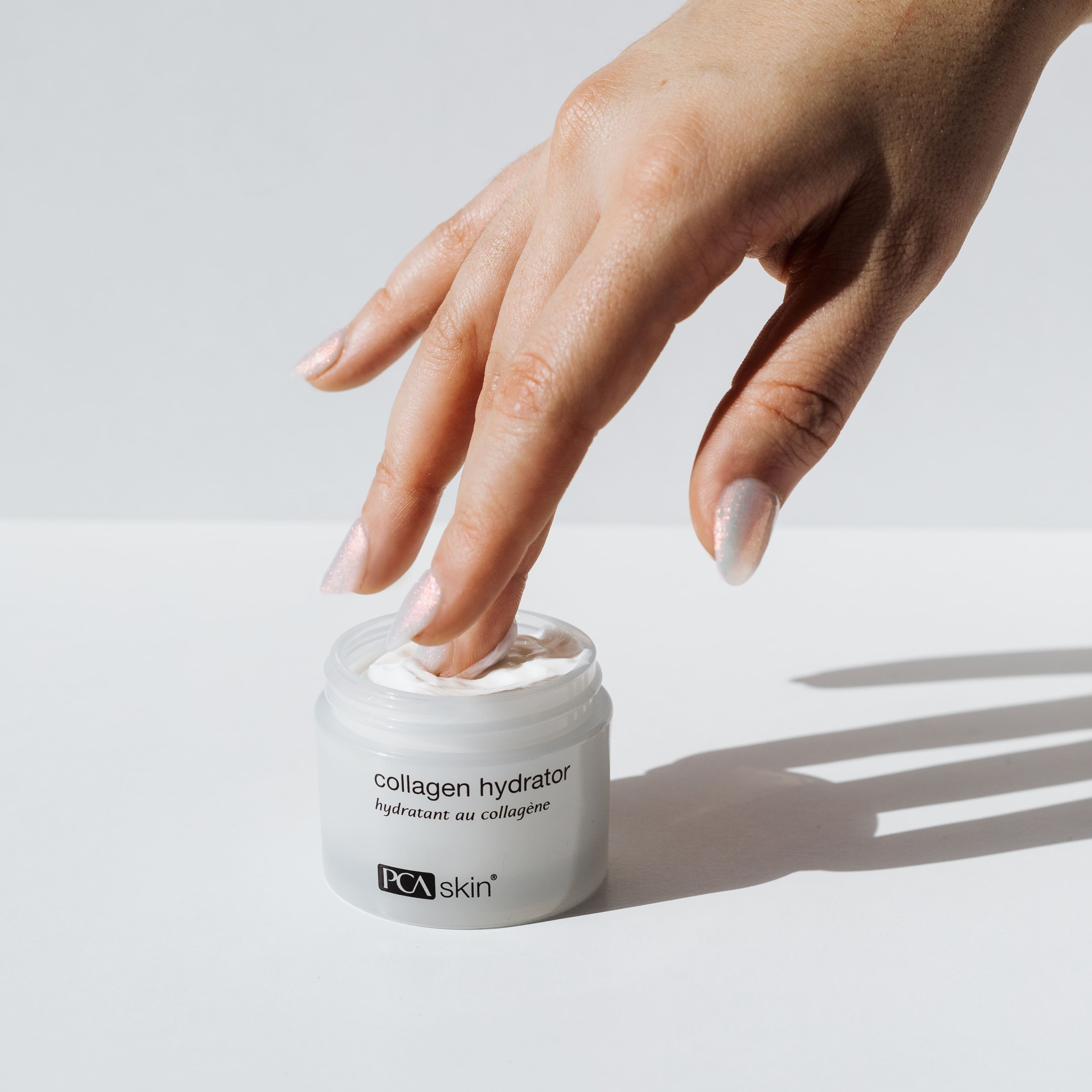 A hand with manicured nails dips a finger into an open jar of Collagen Hydrator cream against a light background. The product label reads &quot;collagen hydrator&quot; and &quot;PCA skin.&quot; Providing intense hydration, it&