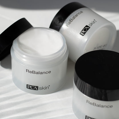 A close-up image of two open jars of ReBalance by PCA Skin. One jar has its lid partially off, revealing the creamy white product inside that offers antioxidant protection for sensitive skin, while the other jar remains sealed. Both jars are placed on a white surface with a soft shadow in the background.