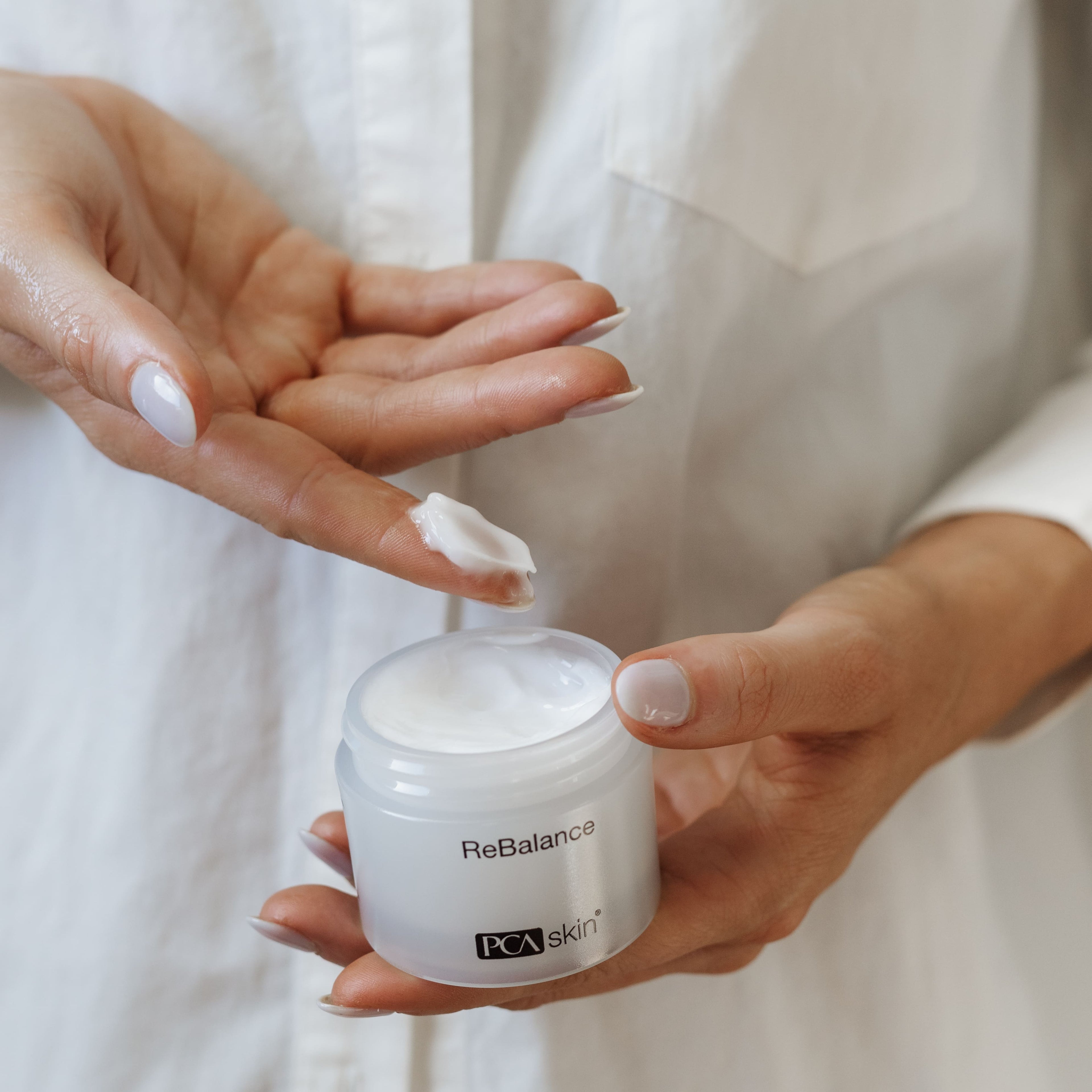 A person in a white shirt is holding an open jar of ReBalance in one hand and applying a small amount to the fingers of the other hand. The jar has a white label with black text. Perfect for sensitive skin, this moisturising cream also offers antioxidant protection.