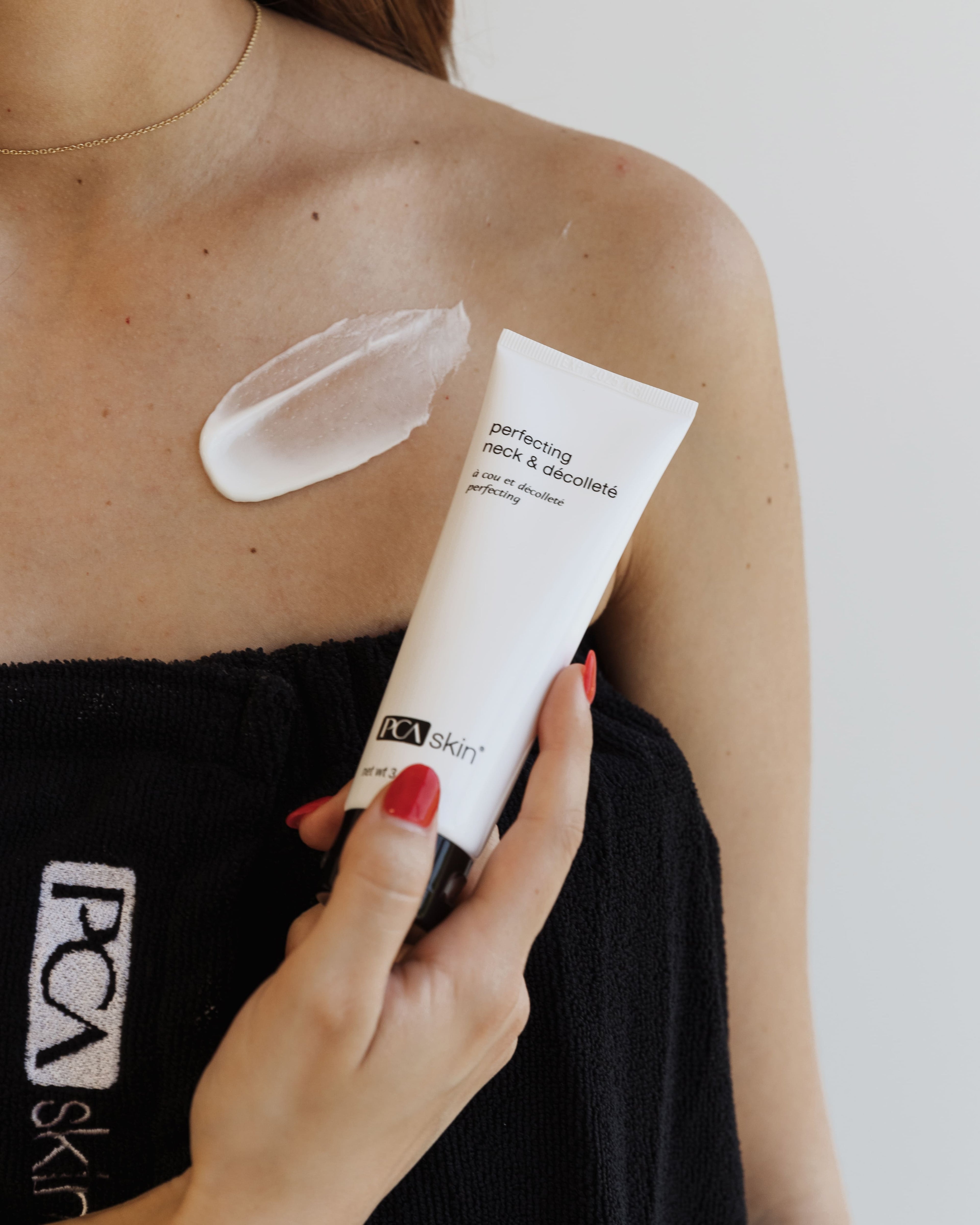 A close-up of a person holding a tube of Perfecting Neck &amp; Décolleté cream with a small amount applied to their upper chest, targeting fine lines. The person is wearing a black towel with the PCA Skin logo.