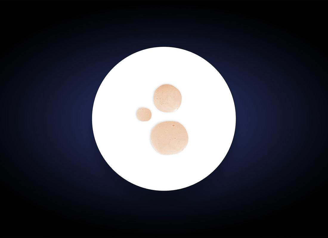 A white plate with three beige sauce droplets of varying sizes sits against a dark, gradient background. The droplets, reminiscent of a luxurious Hydrating Toner, are arranged with the smallest on the left, the medium-sized in the top right, and the largest in the bottom right—offering both hydration and antioxidant protection.