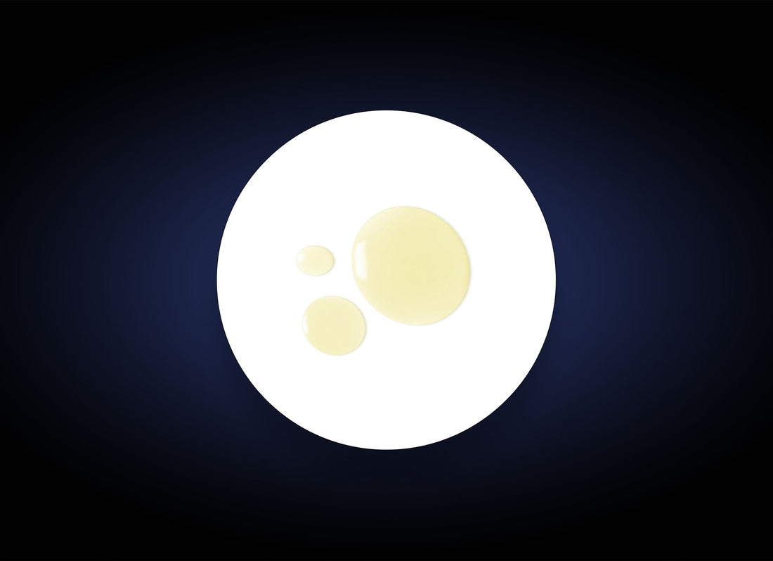 An image shows a dark blue background with an oval white shape in the center. Inside the white oval, there are three irregularly-sized transparent yellow droplets clustered together, reminiscent of Daily Cleansing Oil Makeup Remover used for gentle skin hydration.