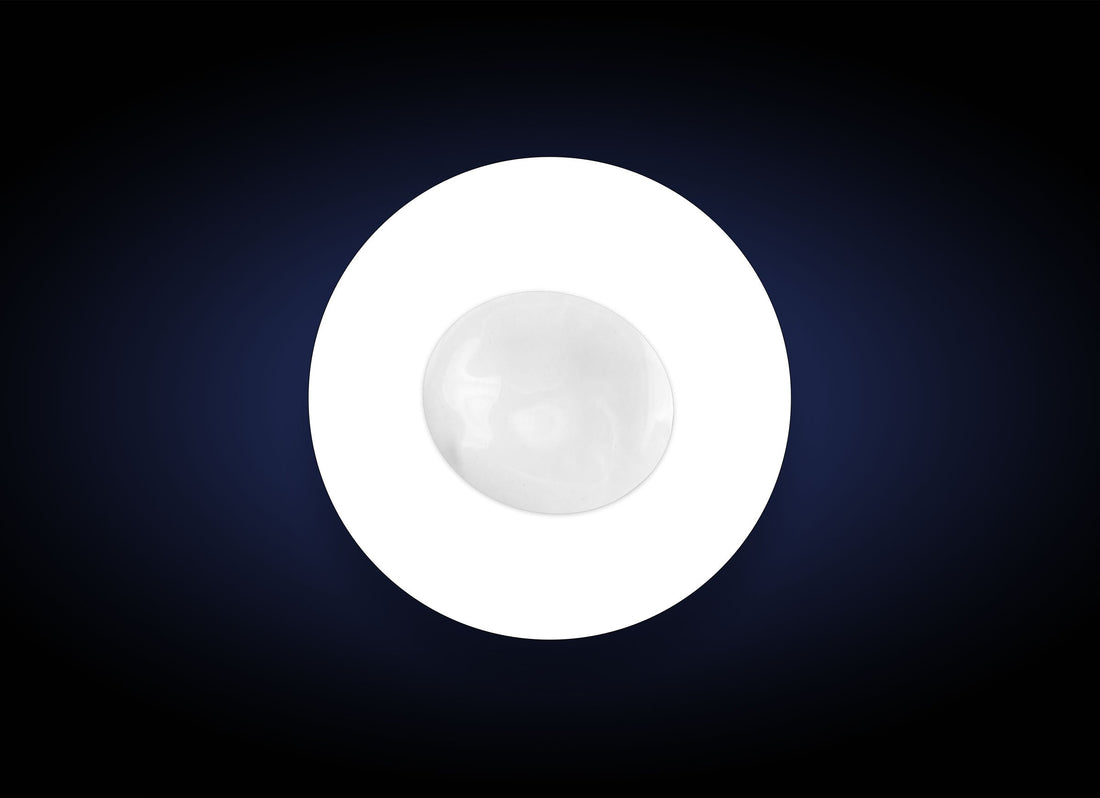 A single white round object, resembling a marble or a smooth stone, placed in the center of a white circle against a dark, gradient background. The circle and background create a striking contrast, emphasizing the simplicity and symmetry of the composition, akin to how Hyaluronic Acid Lip Booster highlights natural beauty.