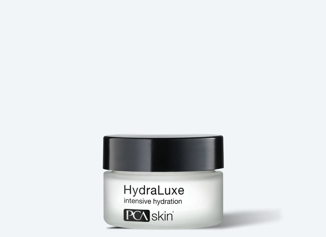 A product image of a frosted glass jar with a black lid, labeled "HydraLuxe Intensive Hydration" by PCA Skin. The white jar, featuring black text that highlights its ability to deeply hydrate and soften fine lines, is displayed against a light gray background.