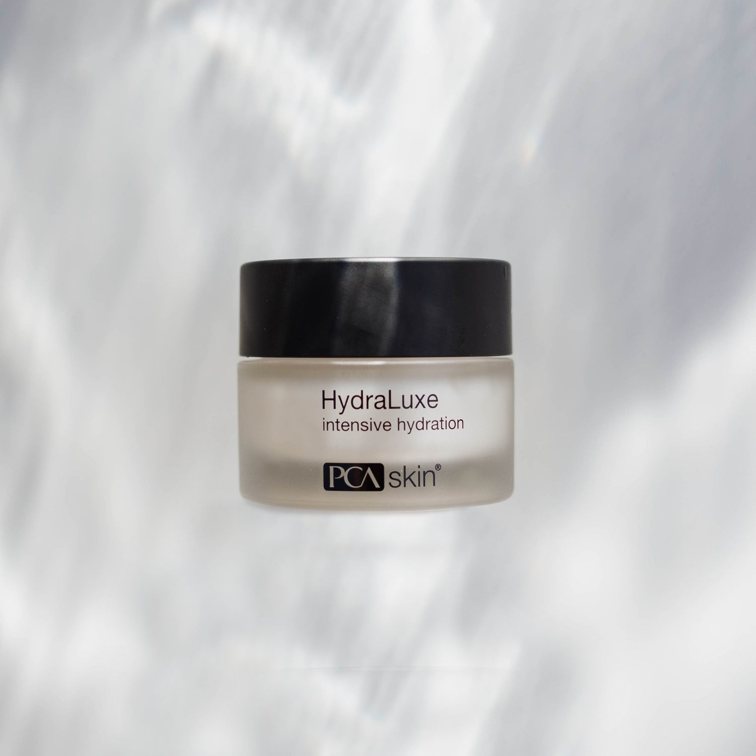 A frosted glass jar with a black lid containing HydraLuxe Intensive Hydration cream. The text on the jar reads &quot;HydraLuxe intensive hydration.&quot; Providing deep hydration, this cream also softens fine lines. The background is light with subtle blurred patterns.