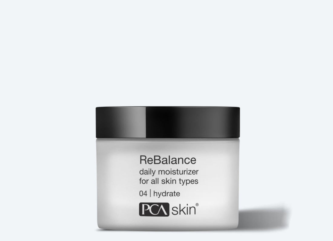 A jar of ReBalance, a daily moisturizer suitable for all skin types, including sensitive skin. The white jar with a black lid features a label that reads &quot;ReBalance daily moisturizer for all skin types 04 | hydrate&quot; and the &quot;PCA Skin&quot; logo at the bottom. The plain light gray background highlights its sleek design.