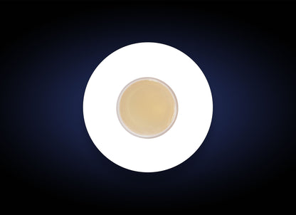 A top-down view of a cup filled with a light beige liquid, possibly tea or coffee, placed on a white circle against a dark background. The contrast between the white circle and dark backdrop highlights the cup centrally, akin to how the Blemish Control Bar targets acne breakouts on oily skin.