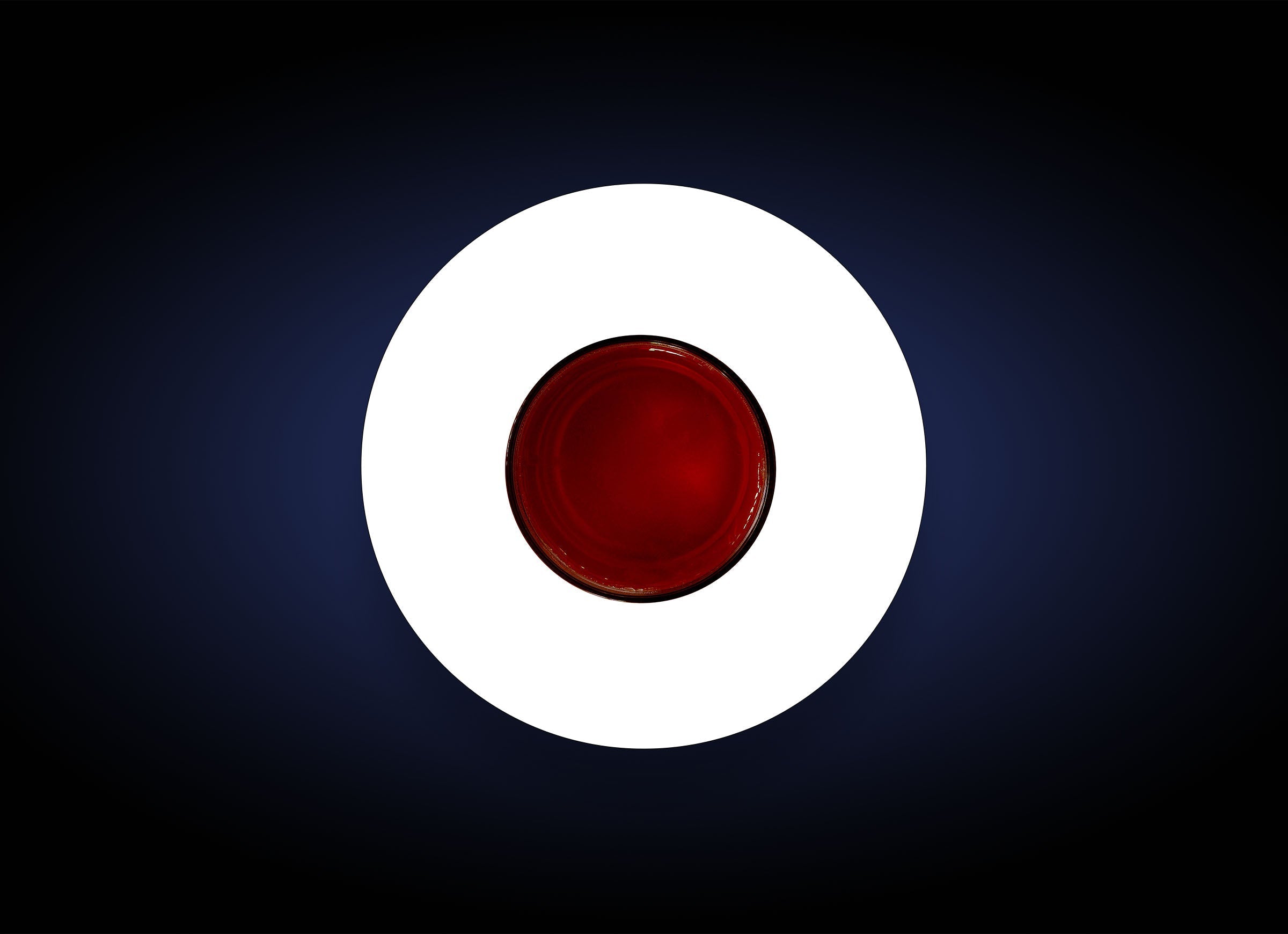 Aerial view of a round, red object centered on a dark blue background with a white circular border around it. The red object, resembling a glossy Pigment Bar, creates a striking visual effect with its vibrant color contrast and polished finish.