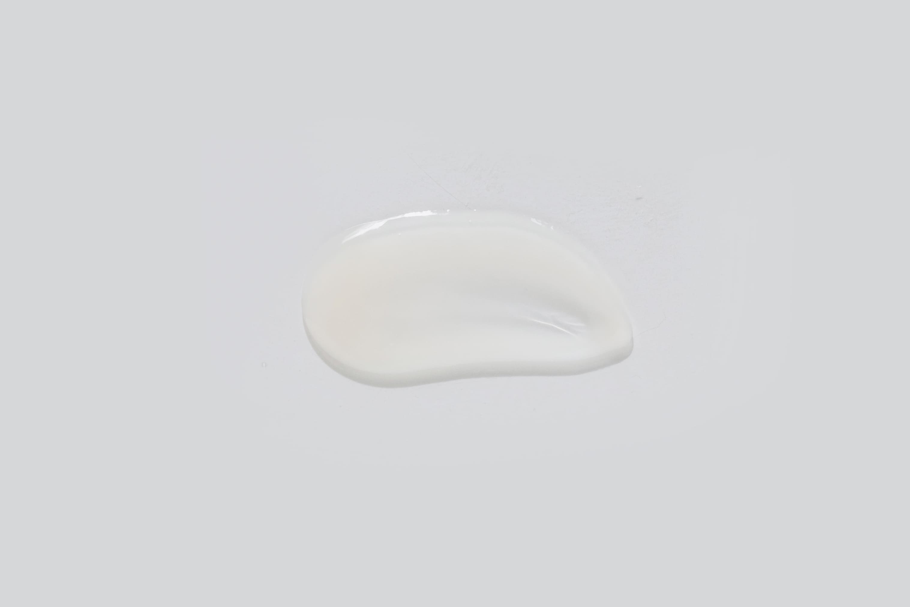 A small puddle of Rejuvenating Serum, rich with anti-aging ingredients, spilled on a light grey surface. The texture appears smooth and slightly glossy.