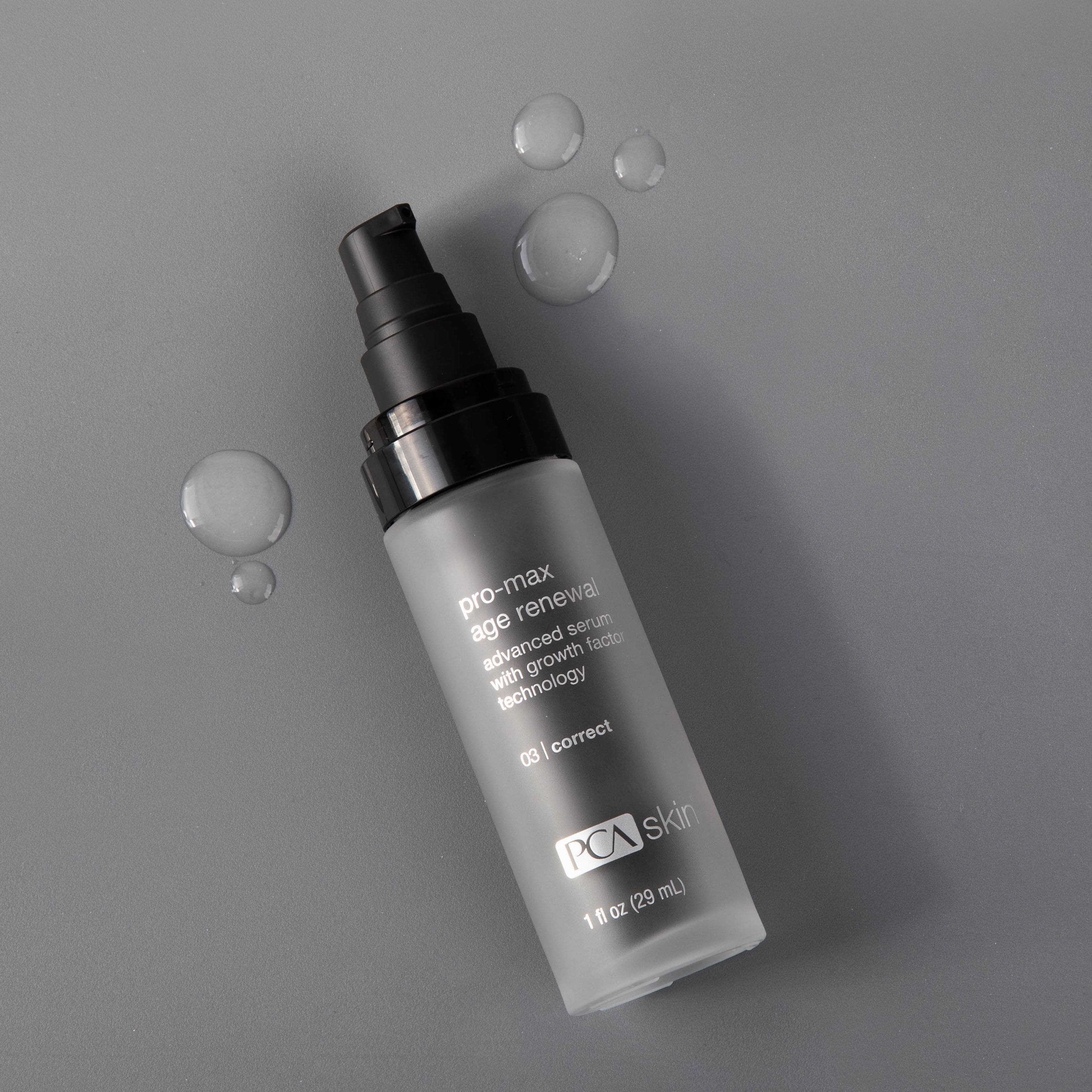 A bottle of Pro-Max Age Renewal serum is lying on a gray surface. The cylindrical bottle boasts a black pump top, with the product description clearly visible. A few droplets, enriched with Micro Growth Factor Technology to boost collagen production, are scattered around it.