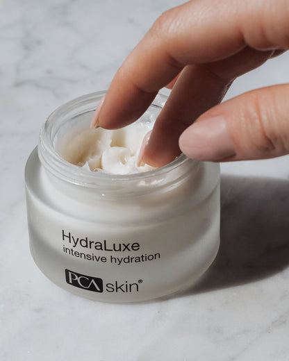 A hand dips two fingers into an open jar of HydraLuxe Intensive Hydration by PCA Skin, resting on a marble surface. The smooth, white cream promises deep hydration and softens fine lines, suggesting a rich texture perfect for moisturizing purposes.