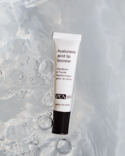 A tube of Hyaluronic Acid Lip Booster on a wet, reflective surface with scattered water droplets around it. The tube is white with black text and has the brand &quot;PCA Skin&quot; printed on the bottom, promising to enhance your lips while supporting collagen production.