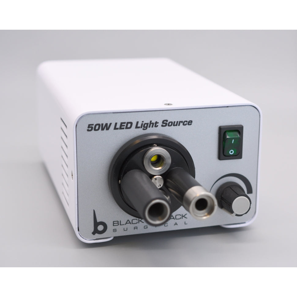 A rectangular white medical device labeled "Onyx-Lite 50Wt LED Light Source w/4-Port Turret" on the front. It features a power switch, two cylindrical ports, and a control knob on the front panel with "Black Jack Surgical" branding at the bottom. The 6500K color temperature ensures precise illumination in a plain grey background.