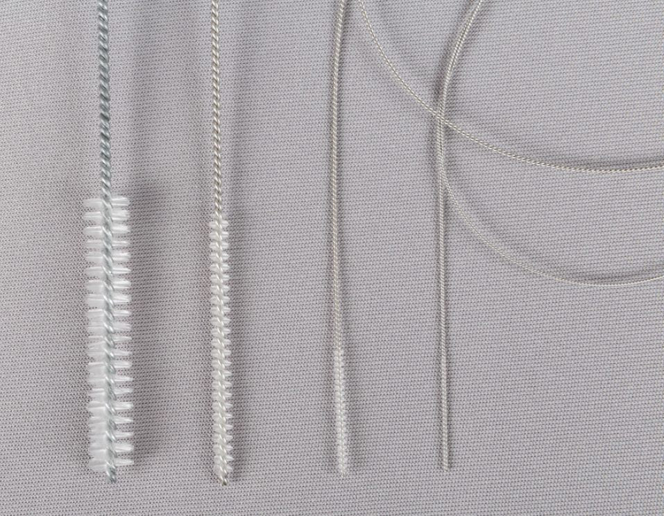 A close-up of four Cannula Cleaning Brushes of varying sizes arranged vertically on a grey textured surface. Each brush has fine bristles and metal handles, with the bristle density and wire thickness differing among them.