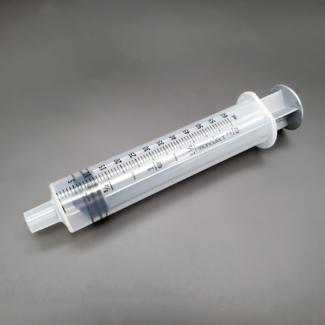 A 60CC Monoject Syringe With Toomey Tip, Sterile - Box of 20, marked with black measurement lines up to 60 ml, lies on a flat, gray surface. The syringe does not have a needle attached to its tip.