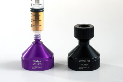 A close-up of two medical devices on a white background. The Decanting Stand™ on the left is purple and labeled &quot;Tulip, ATLLDS, LOT 2796-A,&quot; with a syringe inserted at the top, designed for anaerobic transfers. The Decanting Stand™ on the right is black and labeled &quot;AT60LLDS, LOT 17382-01.