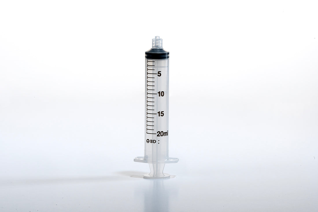 A clear, 20ml syringe is standing upright against a white background. The 20cc Luer BD Syringe, often referred to in the medical field as a 20cc device, is marked with black measurement lines and numbers from 0 to 20. It features a Luer Lock tip and a finger grip at the bottom.