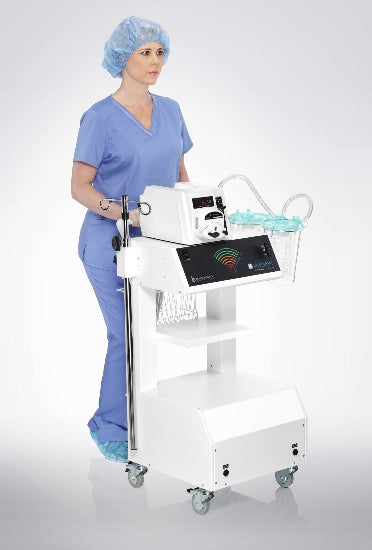 A healthcare professional wearing blue scrubs, a surgical cap, and shoe covers stands behind a medical cart equipped with the Vitruvian Ultimate Aspirator, Dual Motor. The cart has various compartments and is on wheels, suggesting it is designed for mobility and powerful suction tasks.