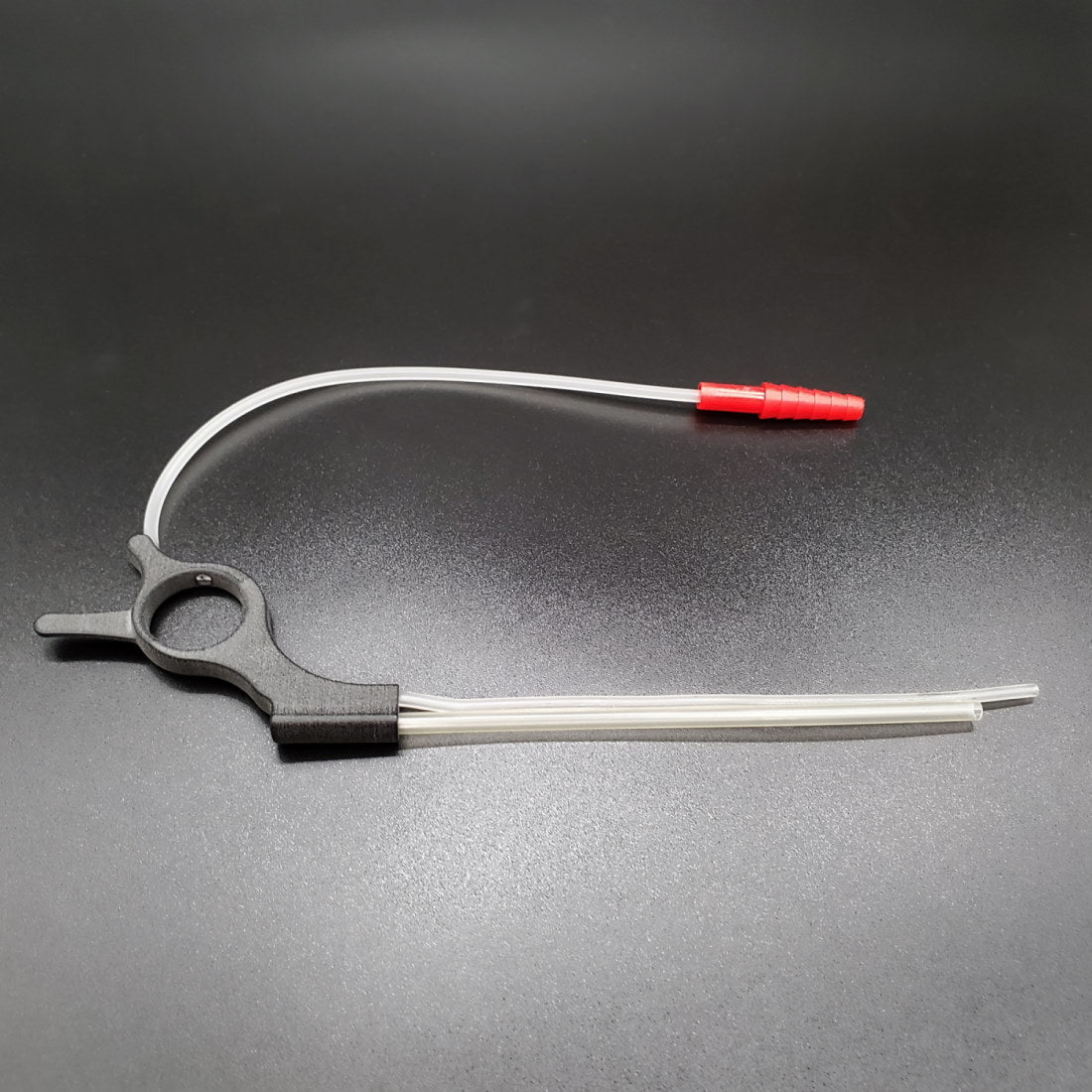 A catheter with a red plug is attached to a MicroTouch™ Sterile Aspiration Tubing - Box of 10 featuring a Ring handle, sitting on a flat, dark surface. The black circular clamp has two extended prongs: one straight and one curved, holding the tubing in place.